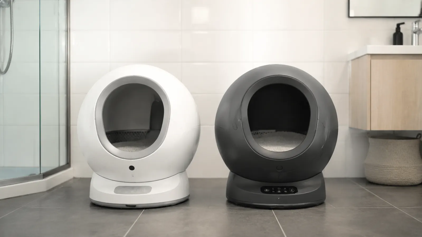 Two self-cleaning litter boxes side by side in a bright modern bathroom, one compact and one globe-shaped